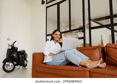 attractive smiling woman sitting on sofa at home cozy relaxed using laptop working remote online wearing white t-shirt and jeans listening to music on earpods - Powered by Shutterstock