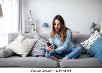 Attraktive lächelnde Frau mit Smartphone, während sie auf dem Sofa zu Hause sitzt. Kommunikationskonzept und Gemütlichkeit.