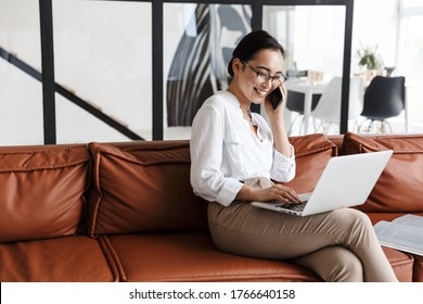 Attractive smiling young asian business woman relaxing on a leather couch at home, working on laptop computer, talking on mobile phone