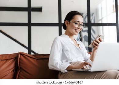 Attractive Smiling Young Asian Business Woman Relaxing On A Leather Couch At Home, Working On Laptop Computer, Holding Mobile Phone