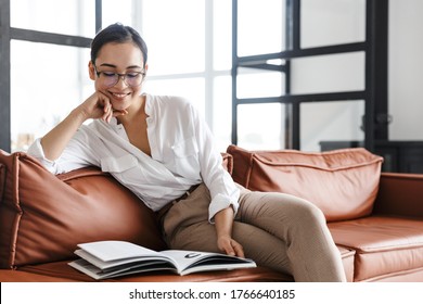 Attractive Smiling Young Asian Business Woman Relaxing On A Leather Couch At Home, Reading A Magazine