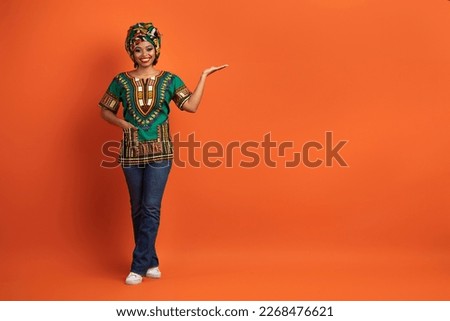 Attractive smiling young black woman in colorful traditional outwear and bright turban pointing at copy space for advertisement over orange studio background, showing nice offer, full length shot