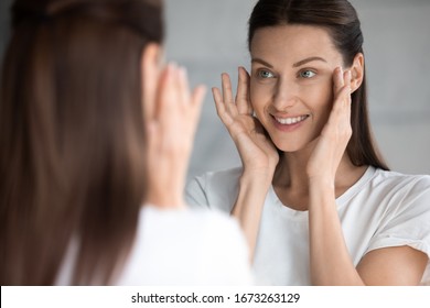 Attractive Smiling Young Brunette Woman Looking At Mirror, Touching Face. Happy 30s Lady Satisfied With Moisturized Soft Skin Condition After Professional Cosmetology Services, Skincare Concept.