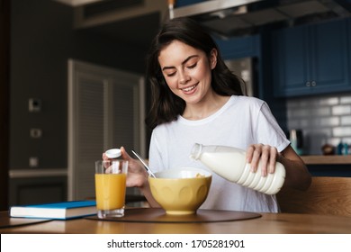 Attractive smiling young brunette woman having a healthy breakfast while sitting at the kitchen table, pouring milk in a cereal - Powered by Shutterstock