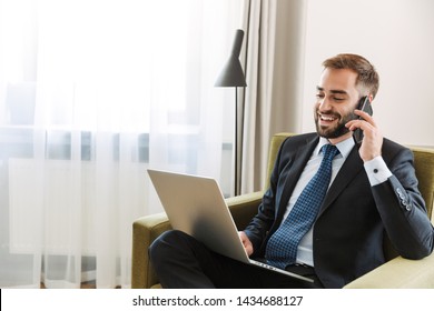 Attractive Smiling Young Businessman Wearing Suit Sitting In A Chair At The Hotel Room, Working On Laptop Computer