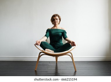 Attractive smiling young Caucasian woman in stylish jumpsuit sitting on chair in lotus pose in spacious empty room keeping eyes closed, meditating, practising yoga, feeling peaceful and relaxed.