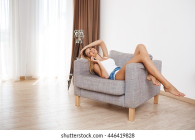 Attractive smiling young girl lying on armchair and talking on phone  at living room