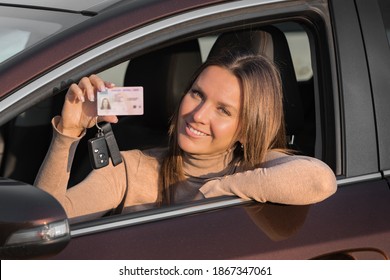 Attractive smiling young woman proudly showing her driving license out of car window. Woman has got driving license and feels very happy and excited. Ready to drive