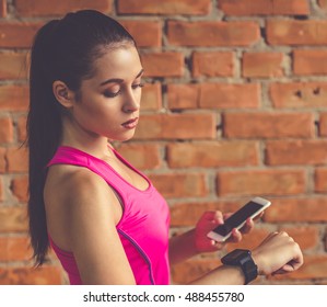 Attractive sports girl is using a smartphone and looking at her fitbit, on brick wall background