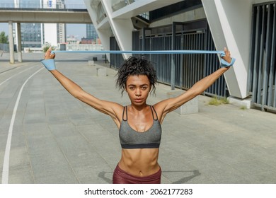 Attractive sportswoman in tracksuit does dynamic lunges raising hands with elastic band near modern building on sunny street - Powered by Shutterstock