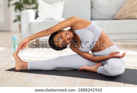 Attractive sporty black woman doing warming up stretching exercses on yoga mat in her home gym. Positive millennial lady bending to her leg, working out muscles flexibility