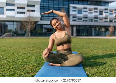 Attractive Sporty Black Woman Sitting On Yoga Mat, Doing Stretching Exercses At Urban Park, Empty Space. Positive African American Female Working Out Muscles Flexibility Outside