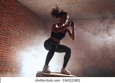 Attractive sporty female with long hair doing squats on wooden box. Process of jumping. Brickwall background