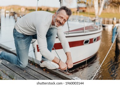Attractive sporty man tying up the mooring rope of his yacht to a wooden jetty in a marina looking up at the camera with a friendly smile