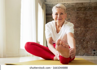 Attractive Sporty Retired Woman Sitting On Floor With One Leg Straight Reaching Forward By Bending At Waist, Trying To Touch Toes. Flexible Mature Female Doing Hamstring Stretch During Yoga Practice