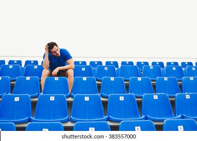 Attractive sporty young man model in blue shirt sitting on blue stadium seats after training staring at field tired and bored. Front view on stadium chairs. Toned whites. Copy space - Powered by Shutterstock