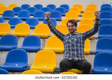 Attractive sporty young man model in blue shirt sitting on blue stadium seats after training staring at field. Perspective view on stadium chairs. Toned whites. Copy space - Shutterstock ID 617201102
