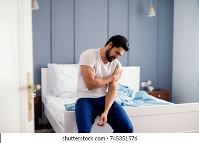 Attractive strong man sitting on the bed and holding painful shoulder with another hand.