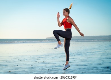 Attractive Strong Young Fit Athletic Blonde Pony Tail Woman Working Out Jumping on Beach Sand in Water Ocean Wearing Red Shirt Yoga Pants at Sunset with Blue Sky Background and Rocks