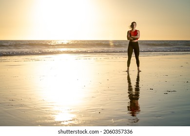 Attractive Strong Young Fit Athletic Blonde Woman In Yoga Pants Arms Crossed Reflection In Water Wearing Red Shirt Yoga Pants On Beach At Sunset With Yellow Sky Background