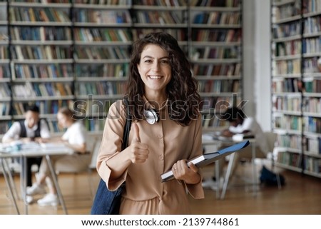 Attractive student 18s girl standing in library showing thumbs up, enjoy interesting effective studies in higher institution. Successful excellent learner, scholarship, good quality education concept