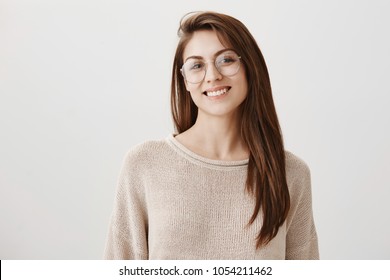 Attractive Student Answering During Lecture. Portrait Of Charming Young Woman In Transparent Glasses Smiling Broadly And Tilting Head, Talking About University Or Part-time Job Over Gray Background