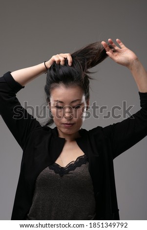 Attractive stylish Asian woman playing with her long hair holding it up in a ponytail above her head over a grey studio background