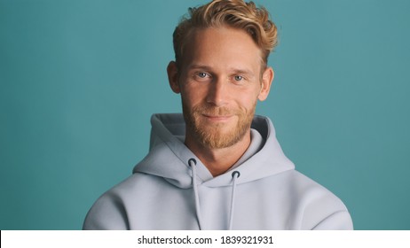 Attractive stylish blond bearded guy in hoodie intently looking in camera and smiling isolated on blue background - Powered by Shutterstock