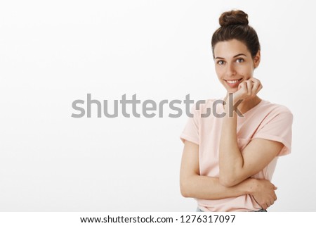 Attractive stylish and confident flirty european woman with cute hair bun in t-shirt smiling joyfully and tender at camera as holding hand on jawline posing and gazing at camera as seducing