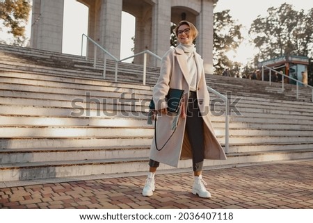 attractive stylish fashionable woman walking in street in elegant style coat wearing glasses, bag and white boots, business lady style, winter autumn spring style fashion