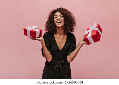 Attractive stylish girl with curly cool hairstyle in polka dot dark clothes laughing, looking into camera and holding gift boxes.