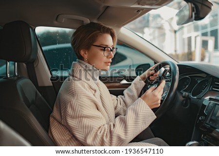 attractive stylish woman sitting in car dressed in coat winter style and glasses using smart phone, business woman busy driving
