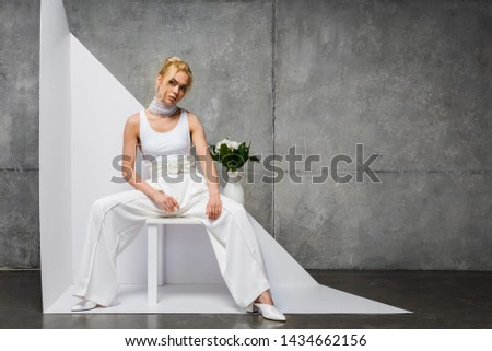 attractive and stylish woman sitting near vase with flowers on white and grey 