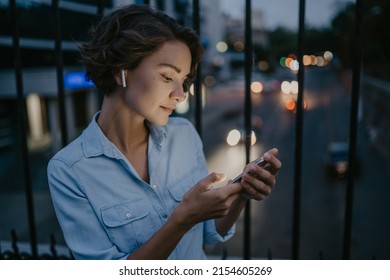 attractive stylish woman walking in street in dusk twilight city in denim shirt, using smartphone, light on face smiling happy, talking on earpods listening to music on earphones