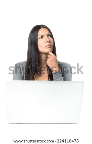 Attractive stylish young businesswoman with a worried frown sitting thinking behind her laptop at her desk staring up into the air with her hand to her chin and a serious expression