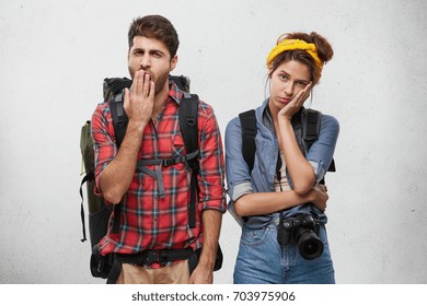 Attractive Stylish Young Couple Of European Travelers Feeling Bored Or Tired: Unshaven Man Covering Mouth While Yawning, His Girlfriend Looking At Camera With Bored Disinterested Expression