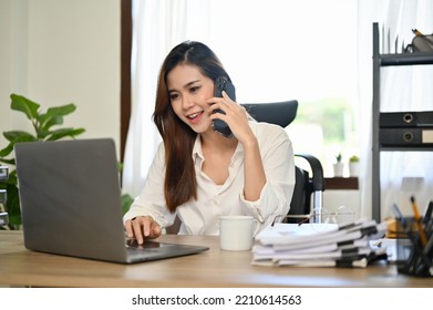 Attractive And Successful Asian Female Boss Or Businesswoman Talking On The Phone With Her Business Partner While Working On Her Project On Laptop Computer In Her Office.