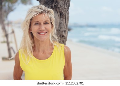 Attractive Tanned Older Blond Woman Standing On A Beach Leaning Against A Tree Grinning Happily At The Camera