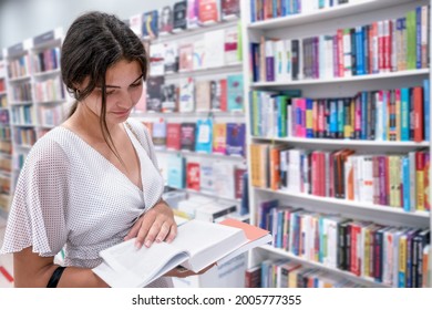 Attractive teenage brunette girl holding and reading book standing in bookstore indoor. Concept studying and education.