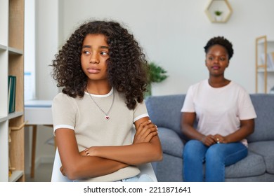 Attractive Teenage Girl Is Looking Sadly Away. Girl Sits With Her Arms Crossed In Closed Pose. Defocused Mom Sitting Behind And Looks At Her Daughter. African American Family At Home.