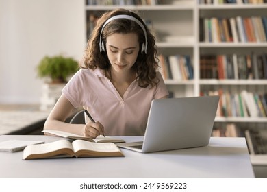 Attractive teenage student girl wear headphones studying in library, sit at desk with laptop, make assignment, writes essay, listen audio course, improve foreign language skills, studying in classroom - Powered by Shutterstock