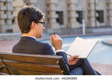 Attractive thoughtful businessman sitting on outdoor bench and writing in blank notepad