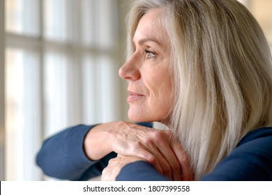 Attractive thoughtful woman with serious expression standing with her hand to her chin staring quietly out of a large window - Powered by Shutterstock