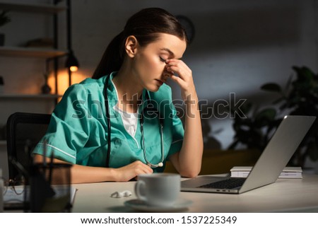 attractive and tired nurse in uniform sitting at table during night shift