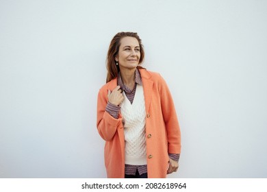 Attractive Trendy Middle Aged Woman Smiling As She Looks Aside With Her Hand To The Lapel Of Her Jacket Against A White Wall With Copyspace