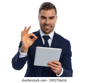 Attractive Unshaved Guy In Navy Blue Suit Holding Tab, Smiling And Making Ok Gesture, Standing Isolated On White Background In Studio