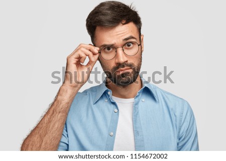 Attractive unshaven male looks curiously through glasses, keeps hand on rim, dressed in fashionable shirt, poses against white background. Young man listens something interesting with wonder