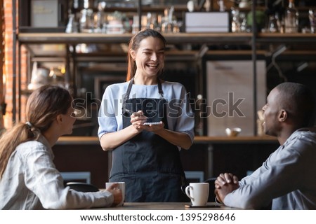 Attractive waitress laughing at African American man funny joke, serving customers, diverse couple making order in cafe, coffeehouse female worker and multiracial visitors having fun