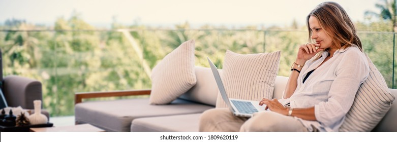 Attractive Woman 40 Years Old In A White Shirt Sitting On A Gray Sofa Working On A Laptop On The Terrace Overlooking The Green Jungle On A Bright Sunny Day.