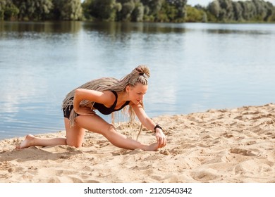 Attractive Woman With Afro Hair Braids Hairstyle Stretching Her Legs Outdoors On The Beach In Summer, Exercising Or Practicing Yoga